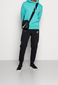 Tyrkysová mikina s logem, černé jogger kalhoty se značkou a černé tenisky. Malá černá crossbody taška má logo.