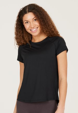 Ung kvinde med krøllet hår smilende, iført en sort t-shirt med korte ærmer og mørke bukser mod en ensfarvet lys baggrund.