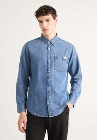 Denim overhemd met knopen in lichtblauw, voorzien van een borstzak, witte knopen en een kraag. Het heeft lange mouwen en een afgeronde onderrand.