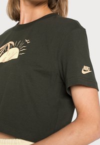 Oliivinvihreä lyhythihainen t-paita, joka on valmistettu puuvillasta ja jossa on beige graafinen suunnittelu auringosta ja linnuista. Nike-logo on painettu hihaan.