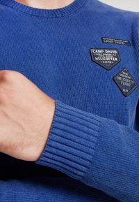 Maglione blu lavorato a maglia con polsini a coste, caratterizzato da diversi pannelli di tessuto che includono loghi e testi in colori a contrasto.