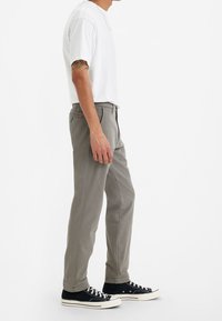 Pantalon en coton gris à coupe droite, poches avant et un petit patch logo rouge, associé à des baskets en toile noires. Chemise blanche à manches courtes.