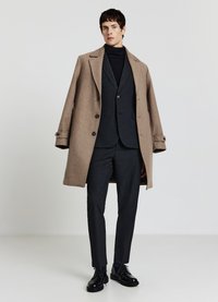 Cappotto lungo di lana marrone indossato sopra un completo scuro e un dolcevita. Vestibilità sartoriale, texture lisce e scarpe nere stringate completano il look.