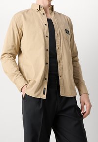 Béžová bavlněná košile na knoflíky s černou logo nášivkou, jednou náprsní kapsou a černými knoflíky, kombinovaná s černými kalhotami. Hladká textura.