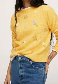 Pull jaune en tricot avec des accents floraux brodés en rose, bleu et blanc, présentant une surface texturée et des poignets côtelés.