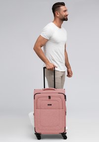 Maleta de ruedas rosa hecha de tela texturizada, con un mango doble, compartimentos con cremallera y acentos negros.