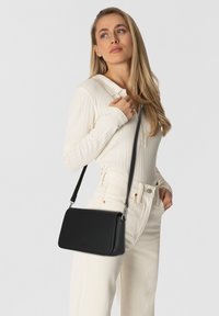 Borsa a tracolla nera in pelle con texture liscia, forma rettangolare e dettagli in metallo argentato, indossata su un completo chiaro.