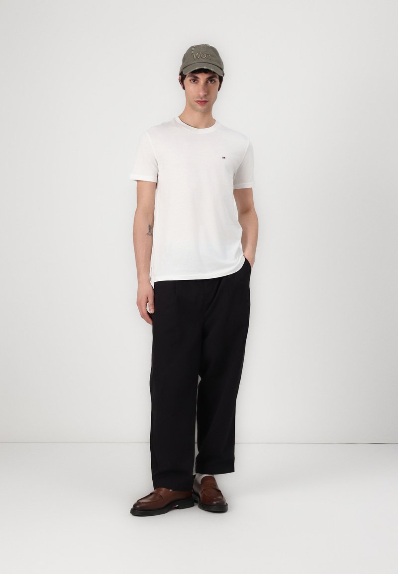 Tricou alb cu guler rotund și un logo mic, pantaloni negri largi și pantofi căprui. Modelul poartă de asemenea o șapcă verde.
