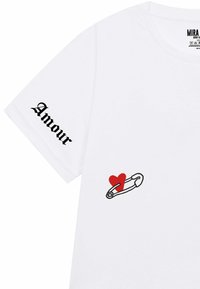 Witte t-shirt met korte mouwen heeft zwarte "Amour" tekst op de linkermouw en een rode hartafbeelding met een veiligheidsspeld op de voorkant.
