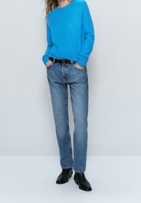 Maglione blu lavorato a maglia abbinato a jeans azzurri chiari, caratterizzato da una vestibilità comoda e una cintura nera. Il modello indossa stivali neri alla caviglia. Sfondo semplice.