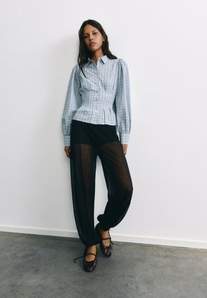 Femme debout contre un mur blanc portant un chemisier ajusté à carreaux, un pantalon noir transparent et des ballerines foncées avec des liens à la cheville.
