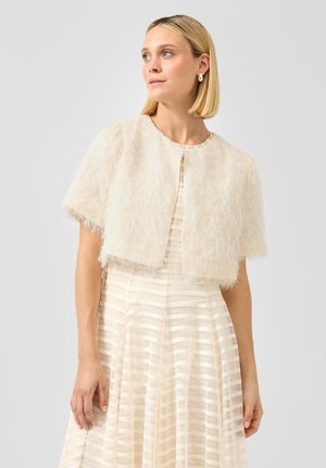 Blonde vrouw die een korte, pluizige crèmekleurige jas draagt over een gestreepte crèmekleurige jurk, en lichtjes opzij kijkt.