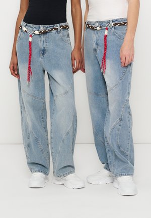 Dvaja ľudia majú na sebe svetlomodré džínsy so širokými nohavicami, biele tenisky, pletené hnedo-biele opasky s červenými strapcami a čierno-biele tričká.