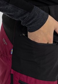 Black and burgundy pants with a zippered pocket, featuring a Swedish flag accent and a logo. Made of a smooth fabric with a fitted design.