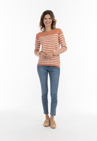 Juostuotas oranžinis ir baltas megztinis su ilgomis rankovėmis, derinamas su šviesiai mėlynomis siaurėjančiomis džinsinėmis kelnėmis ir šviesiais mokasinais. Paprastas iškirpimas ir prigludęs dizainas.