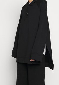 Felpa nera oversize realizzata in tessuto testurizzato. Presenta una tasca a marsupio, cappuccio con cordino e fessure laterali con una vestibilità comoda.