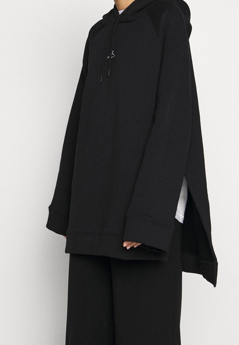 Sudadera negra de gran tamaño hecha de tela texturizada. Cuenta con un bolsillo tipo canguro, capucha con cordones y aberturas laterales, todo con un corte holgado.
