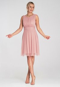 Różowa sukienka bez rękawów z marszczoną górą i dopasowanym pasem; zwiewna spódnica; przezroczysta warstwa; noszona z sandałami na obcasie i naszyjnikiem z warstw.