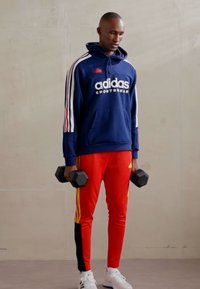 Felpa navy con strisce bianche e logo "adidas SPORTSWEAR", abbinata a pantaloni rossi con accenti gialli, mentre tiene dei manubri neri.