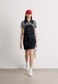 Tumši zila džinsa kopija kleita ar priekšējo kabatu, kas stilizēta virs melnbaltas svītrotas krekla. Baltas čības un sarkans cepure piešķir kopējam tēlam.