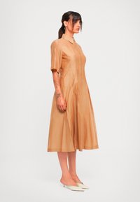Kvinne med mørkt hår i hestehale, iført en beige midikjole med korte ermer og krage, kombinert med hvite spisse kitten heels.