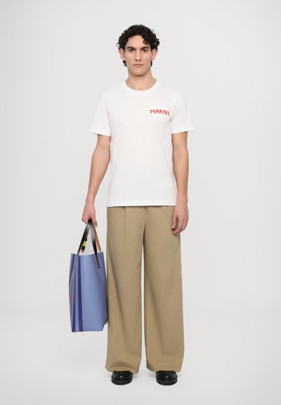 Valkoinen t-paita, jossa on punainen "MARNI"-logo, beige leveät housut ja mustat kengät. Pidättelee sinistä laukkua, jossa on raidallisia yksityiskohtia ja mustat kahvat.
