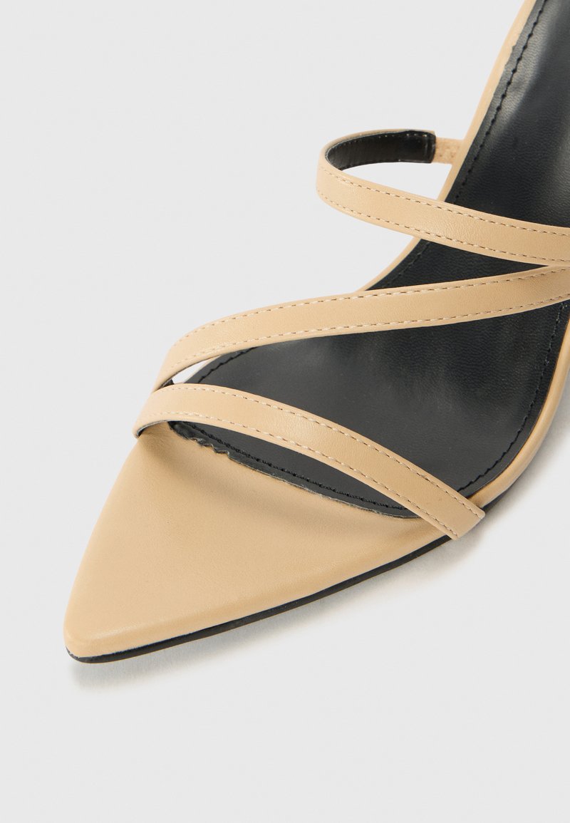 Sandale à talons hauts beige avec un bout pointu et de fines sangles croisées. Semelle intérieure noire et finition en cuir lisse. Pas de matériel ou de motifs visibles.
