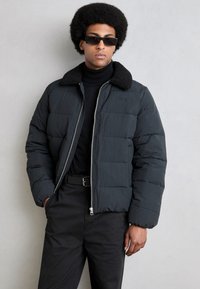 Sort pufferjakke med en struktureret sort krave, lynlås foran og et quiltet design. Båret over en sort turtleneck og parret med sorte bukser.