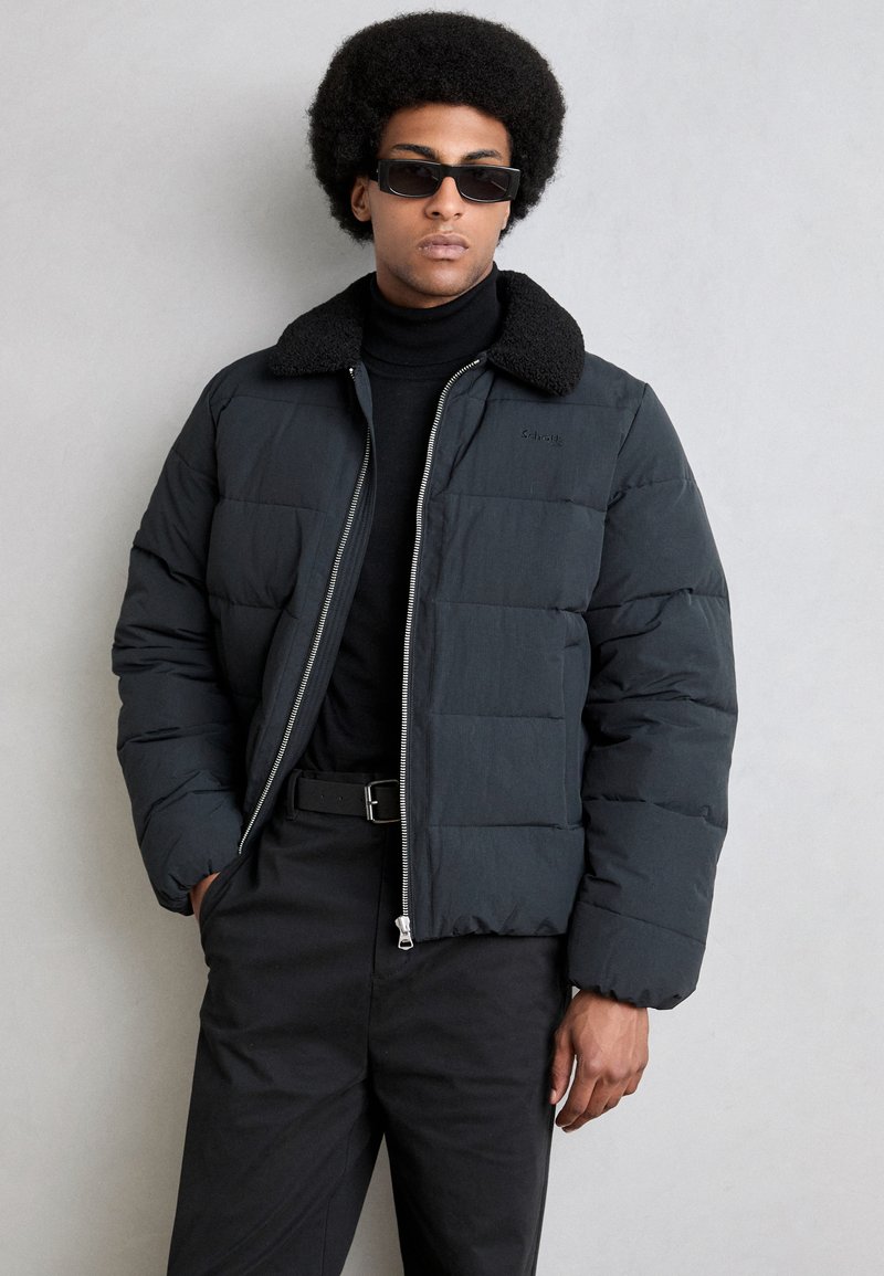 Sort pufferjakke med en struktureret sort krave, lynlås foran og et quiltet design. Båret over en sort turtleneck og parret med sorte bukser.