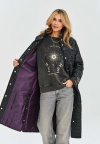 Czarna pikowana długa kurtka z purpurową podszewką, z zatrzaskami. Pod nią ciemny sweter z graficznym nadrukiem i szare dżinsy.