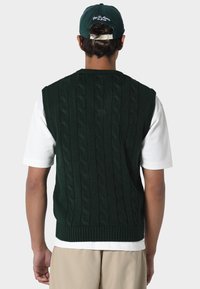 Tummanvihreä neulottu liivi, jossa on palmikkokuvio ja resorireuna, pidetty valkoisen paidan päällä. Tumma lippalakki näkyvissä. Tausta on yksivärinen valkoinen.