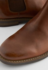 Botas de tobillo de cuero marrón con un acabado suave, paneles laterales elásticos y una costura clara que contrasta en la suela.