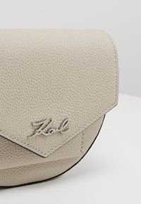 Beige nahkalaukku, jossa on kuvioitu pinta, läppäkiinnitys ja hopea logo. Pyöreä muoto, jossa on kontrastiväriset mustat reunat.