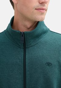 Giacca in pile verde scuro con colletto alto, zip frontale e un logo discreto sul petto; tessuto testurizzato con un design sportivo.