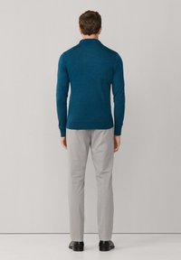 Zielona koszula polo z długim rękawem i kołnierzykiem, w połączeniu z jasnoszarymi spodniami i czarnymi butami, widoczna z tyłu na neutralnym tle.