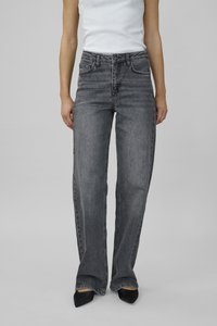 Jeans de mezclilla gris de pierna ancha con cintura alta, bolsillos frontales y un acabado desgastado, combinados con zapatos negros puntiagudos y una camiseta blanca sin mangas.