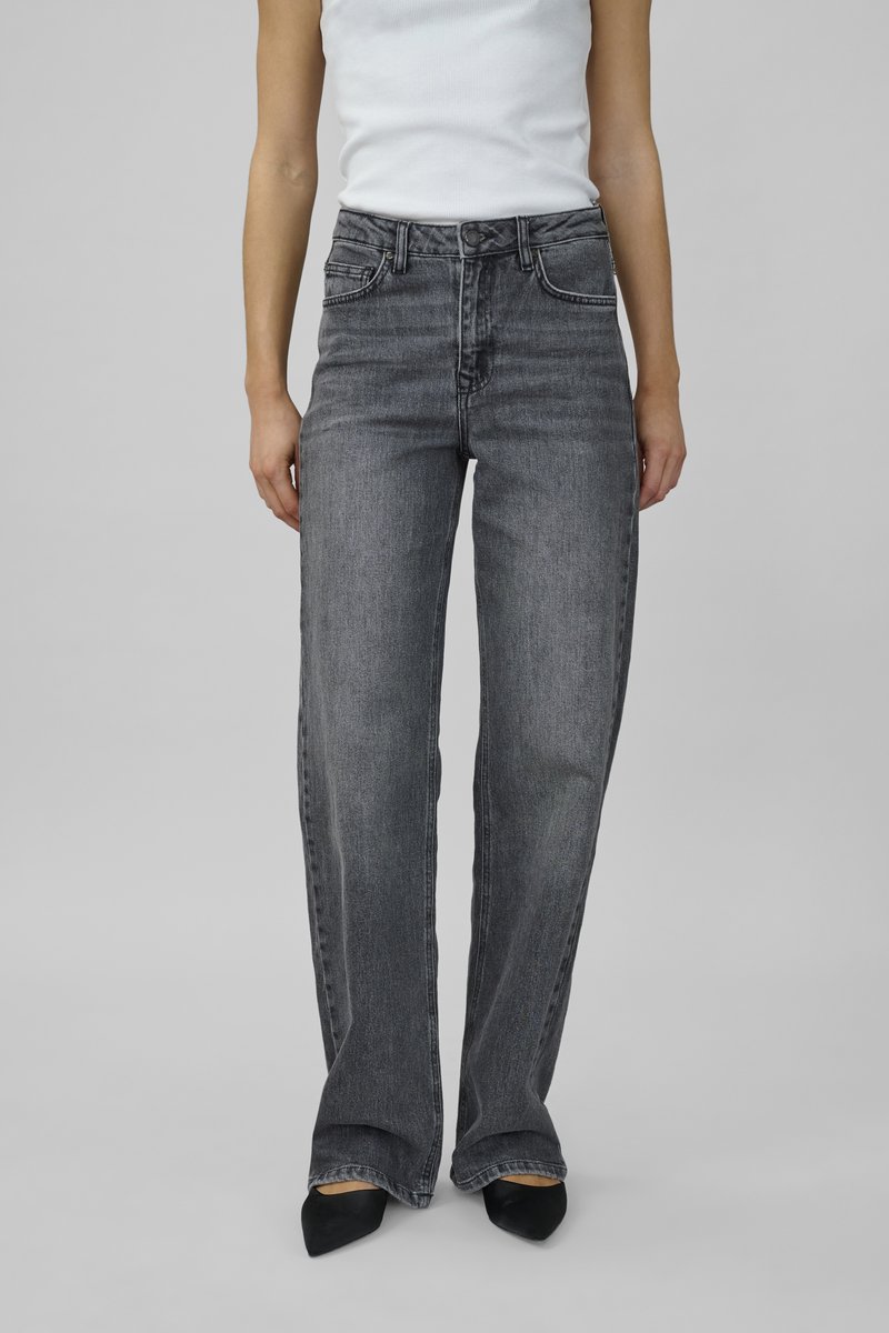 Jeans de mezclilla gris de pierna ancha con cintura alta, bolsillos frontales y un acabado desgastado, combinados con zapatos negros puntiagudos y una camiseta blanca sin mangas.