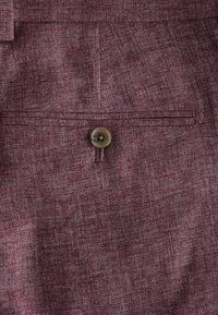 Bordeauxfarbener Stoff mit strukturierter Optik, ausgestattet mit einem einzigen Knopfverschluss und einer kleinen Tasche mit dezentem Stichdetail.