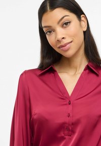 Kvinde med langt mørkt hår iført en dybrød satinbluse med knapper, der smiler blidt mod en ensartet lys baggrund.