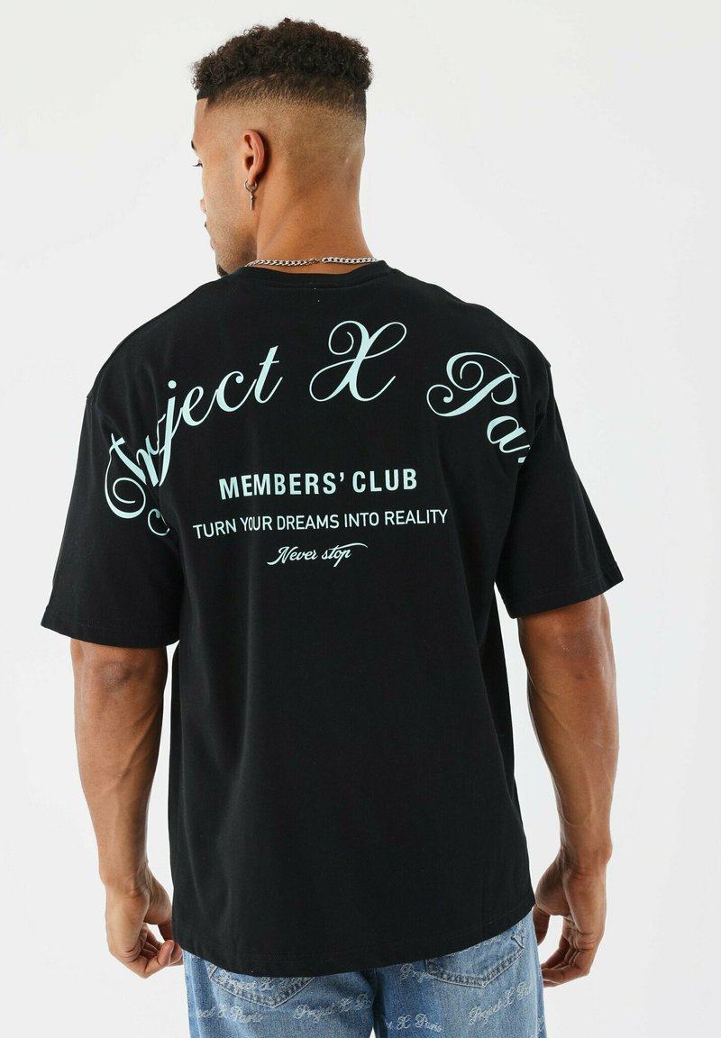 Hombre con cabello rizado corto que lleva una camiseta negra con texto en azul claro "Club de Miembros de Project X Paris, Convierte tus sueños en realidad, Nunca te detengas."