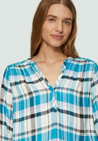 Blau-weiß karierte Bluse mit V-Ausschnitt, in verschiedenen Farbtönen von Türkis, Schwarz und Lavendel. Falten-Details nahe den Schultern.
