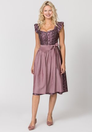 Blonde Frau lächelt, trägt ein violettes Kleid mit floralem Mieder und Rüschenärmeln sowie eine passende einfarbige violette Schürze, die in der Taille gebunden ist.
