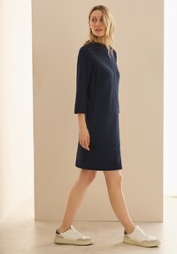 Marineblaues strukturiertes Kleid mit Dreiviertelärmeln, seitlichen Taschen, getragen mit weißen Sneakers mit grauen und burgunderfarbenen Akzenten.