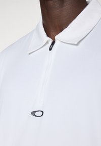 Hvit polokrave skjorte med glidelås, med en liten svart logo på brystet og en jevn, myk tekstur.