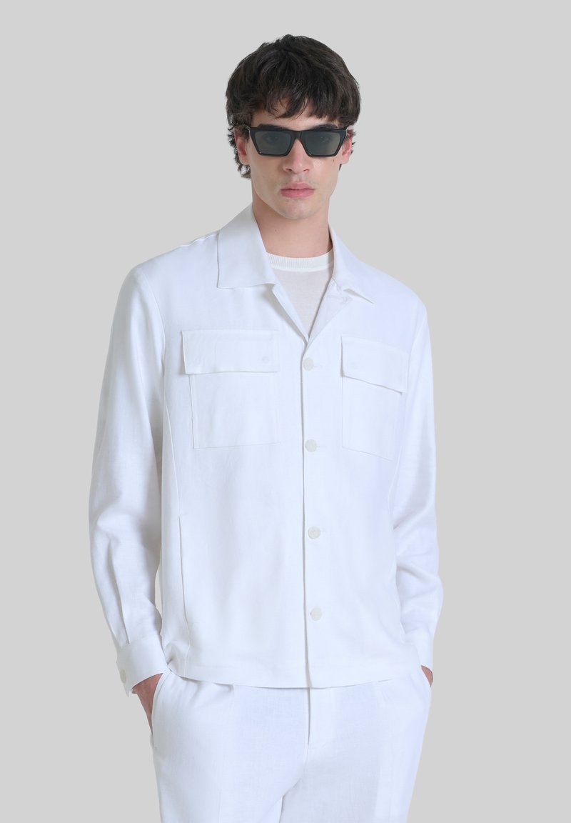Camisa blanca de lino con botones, con dos bolsillos en el pecho, mangas largas y pantalones a juego. Se lleva con gafas de sol rectangulares oscuras.