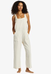 Jumpsuit color crema chiaro realizzato in tessuto testurizzato, con scollatura quadrata, spalline larghe, tasche laterali e un fit rilassato con gambe ampie.