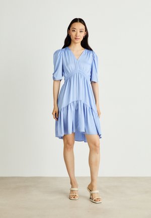 Helles blaues Kleid mit Puffärmeln, V-Ausschnitt und gerafftem Taillenteil. Es hat einen ausgestellten Saum und einen weichen, fließenden Stoff. Trägt dazu beigen Sandalen.