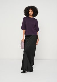 Purpura īss piedurkņu tops kopā ar garu melnu satīna svārku. Modeles rokās ir gaiši violeta soma, uz kājām melnas smailas kurpes.