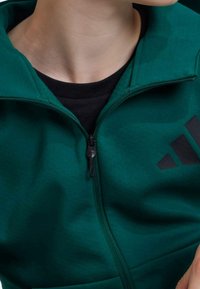 Grön dragkedjejacka av mjukt tyg, med hög krage, svart dragkedja och en svart logotyp med tre ränder på vänster sida.