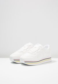 Baskets compensées blanches avec fermeture à lacets et semelles rayées multicolores en rose, noir et jaune sur une surface blanche.